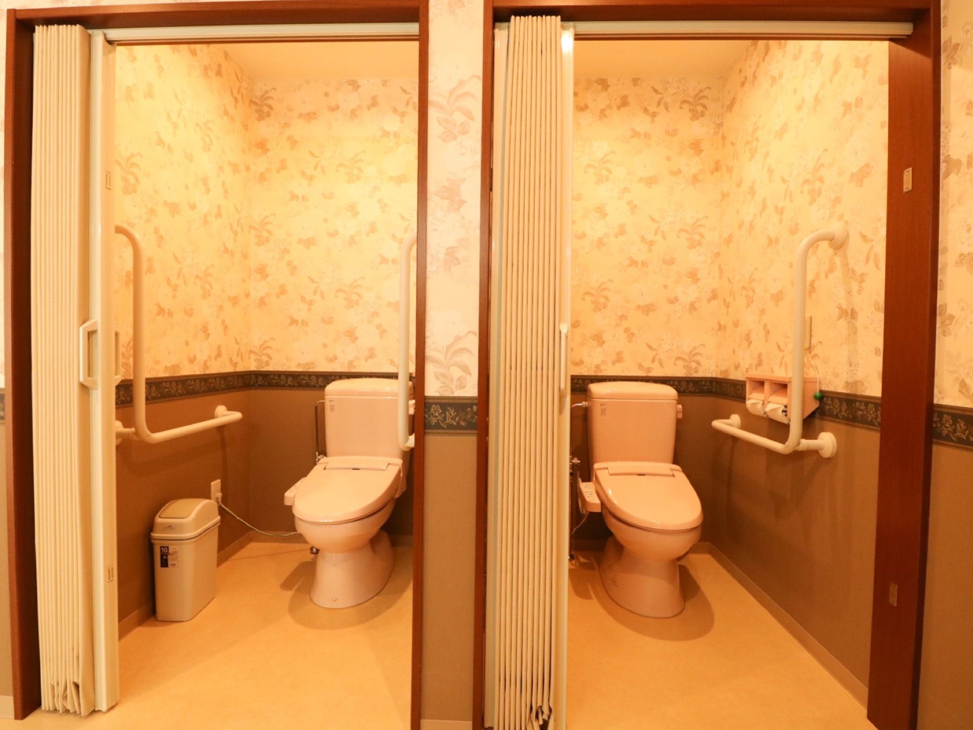 トイレの個室が２ヶ所並んでいる。段差を取り除き、便座の横側の壁にエル型の手すりを設置し、ドアはカーテン式である。