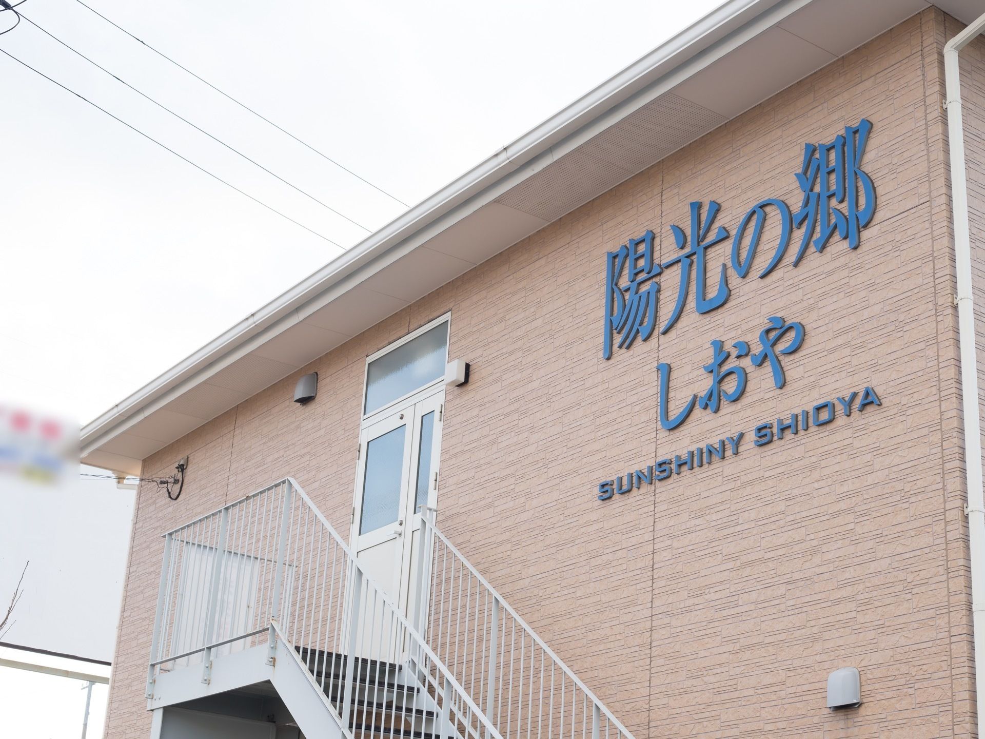 茶色の外壁の屋根近くに、大きな文字で施設名が書かれている。その近くには扉があり、外階段に続いている。