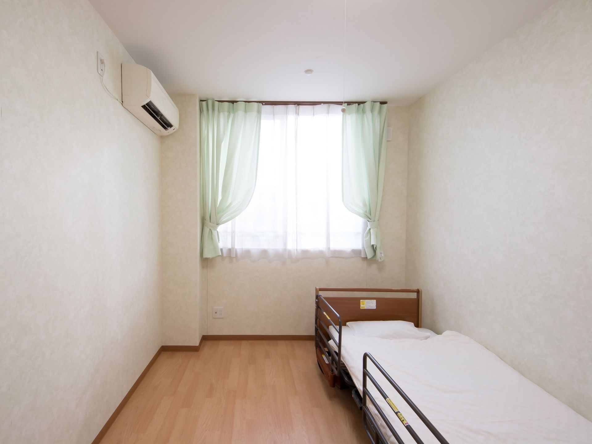 居室の窓には、グリーンのカーテンが掛けられている。エアコンと転落防止の柵がついたベッドが設置されている。