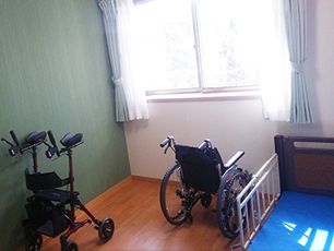 カーテンが付いた大きな窓があるフローリングの部屋には、介護ベッドや車いすなどが置かれ、緑のアクセントクロスが貼られている。