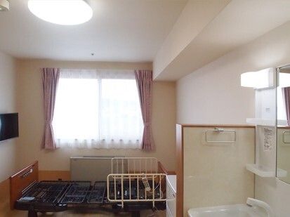 介護ベッドやテレビ、車いす対応の洗面所がある部屋には、レースカーテンとカーテンが付いた大きな窓がある。
