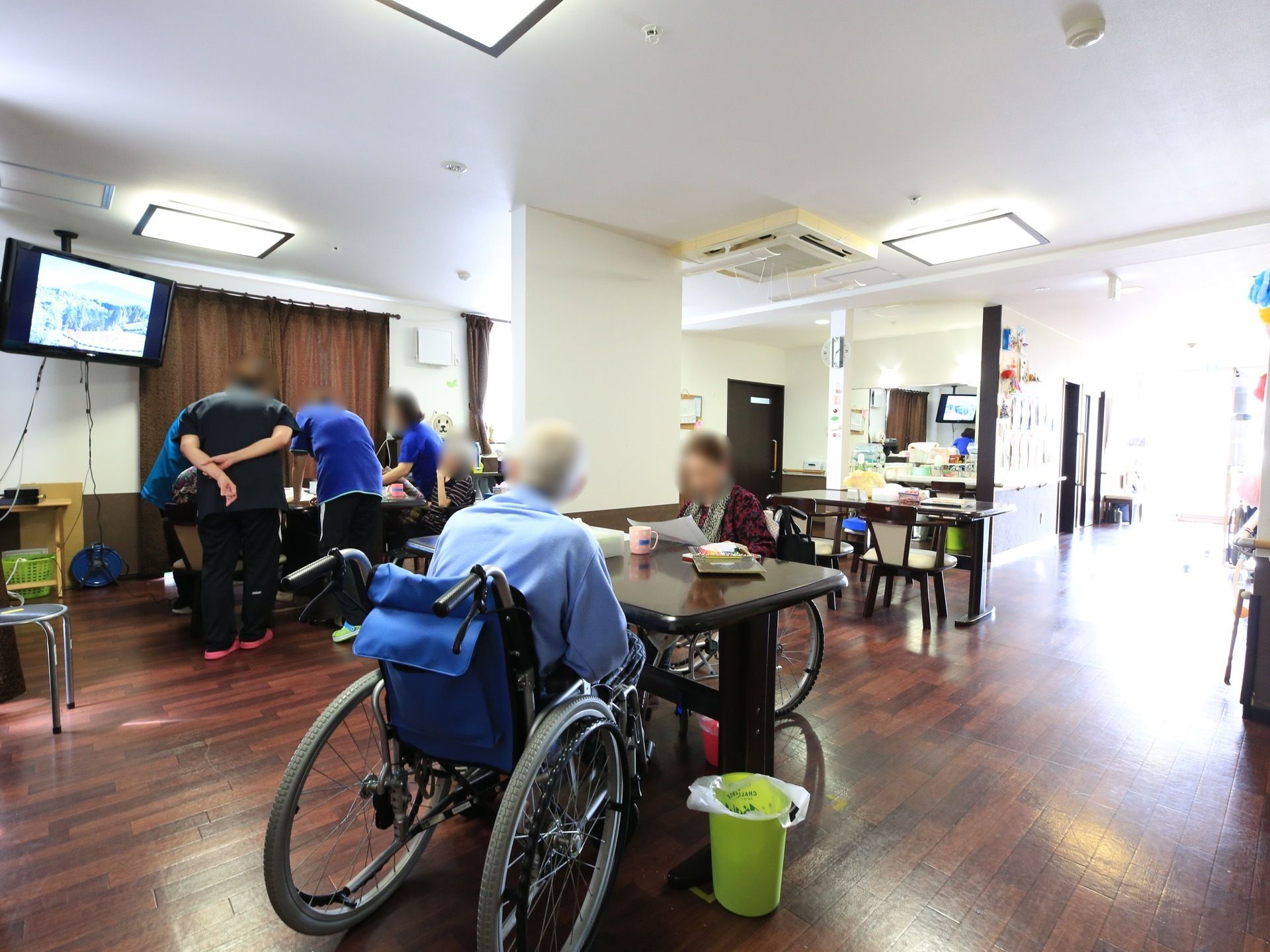 複数のダイニングテーブルが設置されているフロアで、入居者様が車椅子に座ってくつろいでいたり、スタッフが何か話をしたりしている。