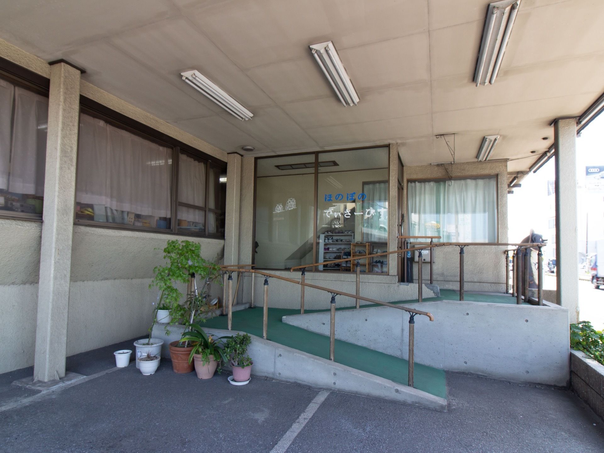 手すり付きのスロープが、玄関の前方に取りつけられている。左端には植木鉢が数個あり、さまざまな草木が植わっている。