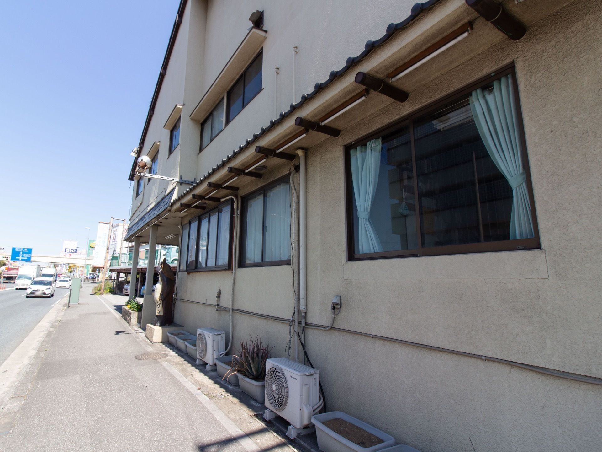 地上２階建ての施設は、アットホームな外観デザインである。左横の道路には、小型トラックなど数多くの車が走行している。