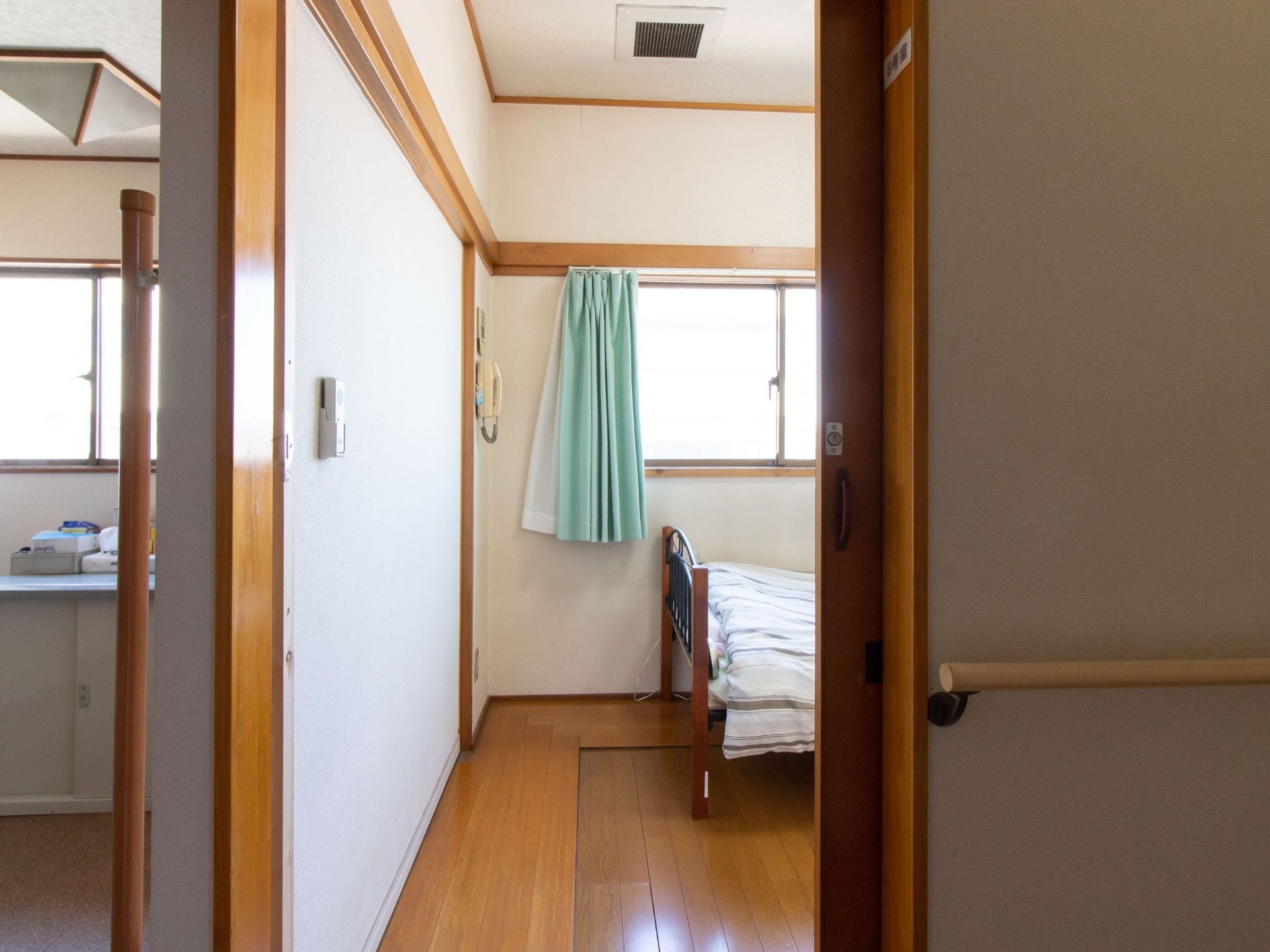 居室の出入口ドアが開けてあり、窓やベッドの一部が見えている。天井には、四角い形の換気口が取りつけられている。
