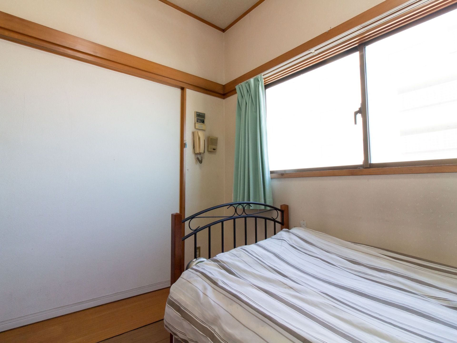 居室の窓際には、おしゃれなアイアンフレームのベッドが置いてある。掛ふとんカバーは、白とグレーのストライプ柄である。