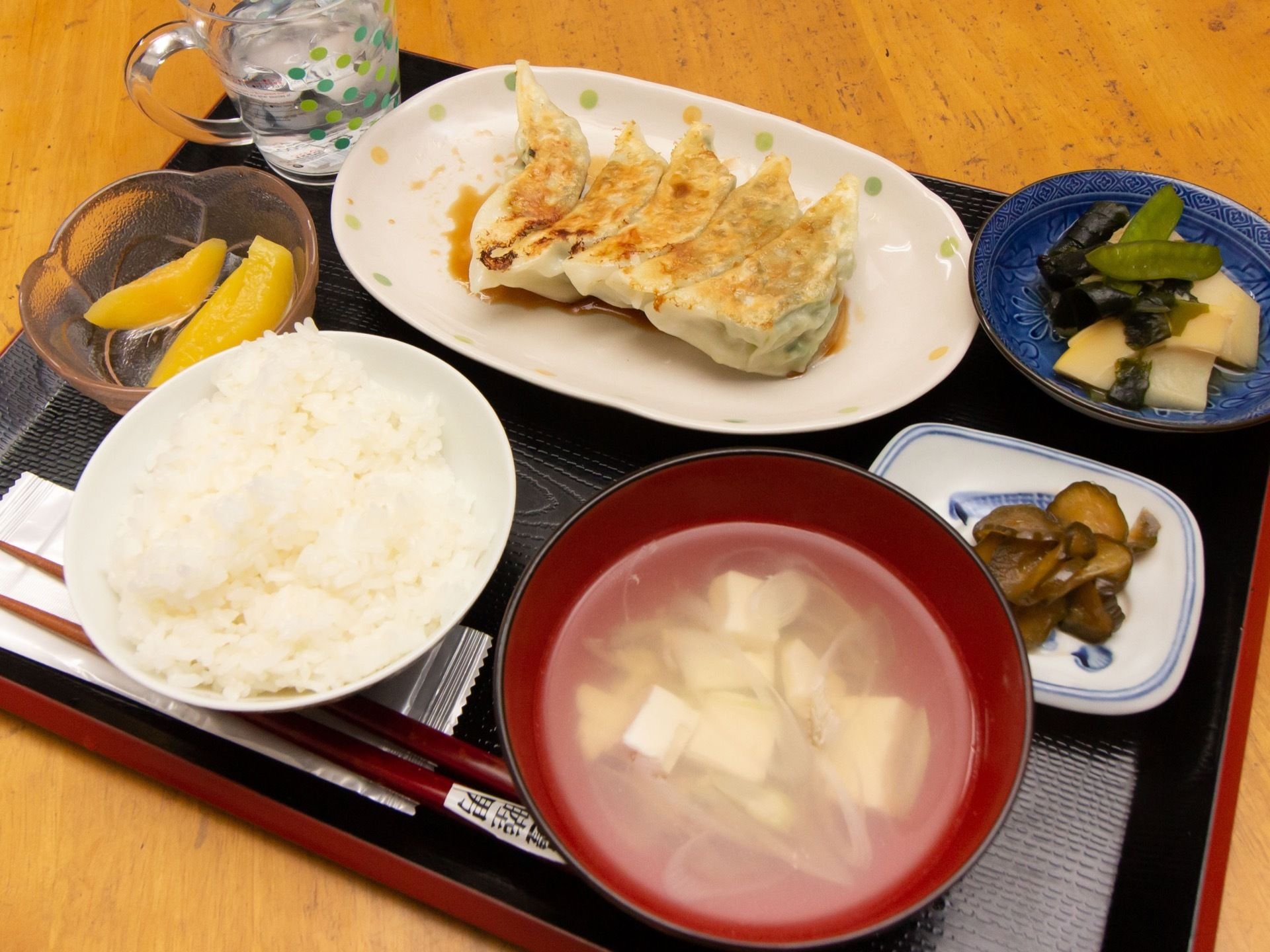 ご飯、おとうふの味噌汁、キュウリの漬物、タケノコの煮物、ギョウザ、デザート用の黄桃などが、お盆にセットされている。