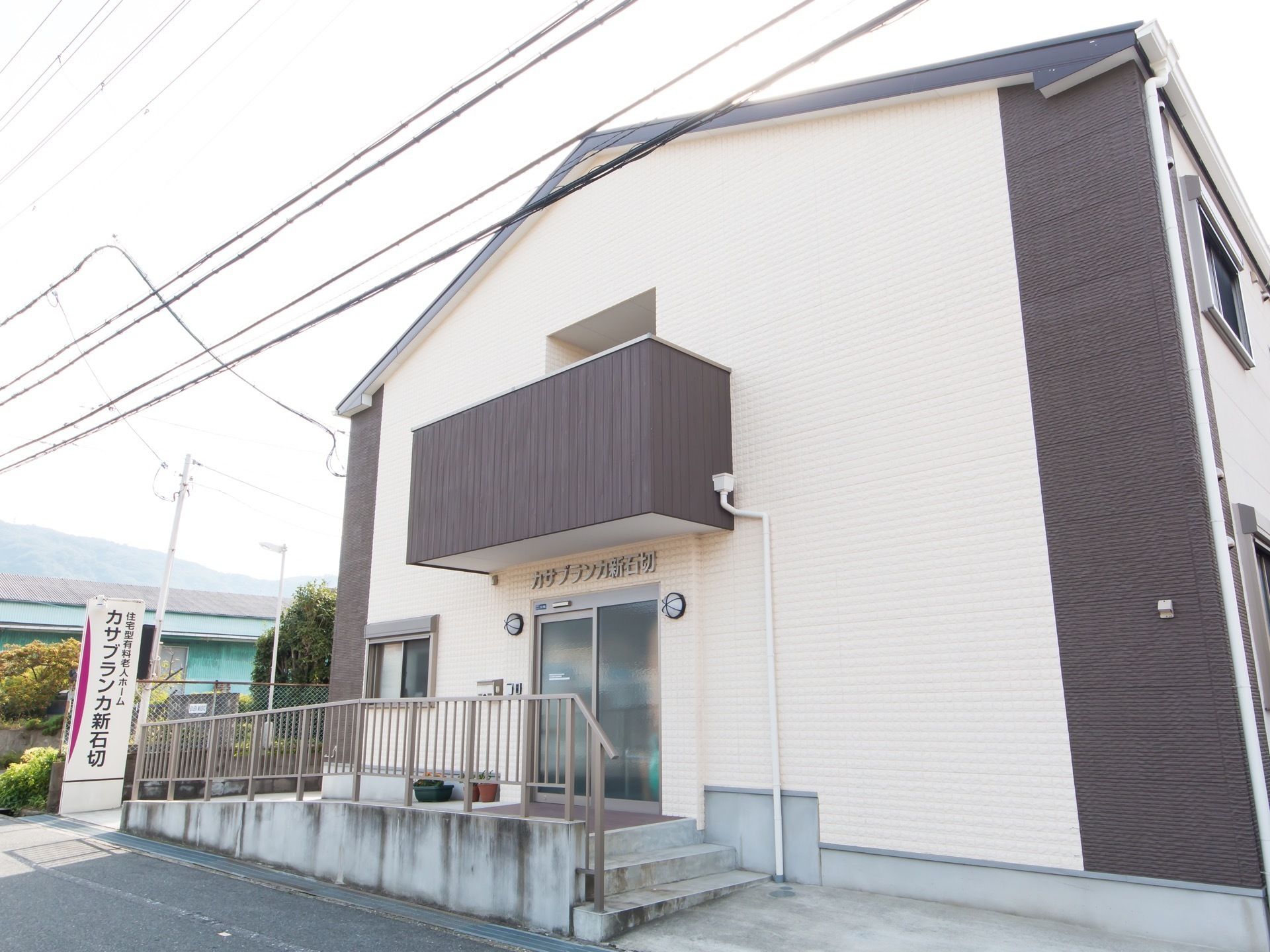 白色と茶色を基調とした２階建ての施設である。道路に面した立地で、敷地境界に縦長の看板が設置されている。