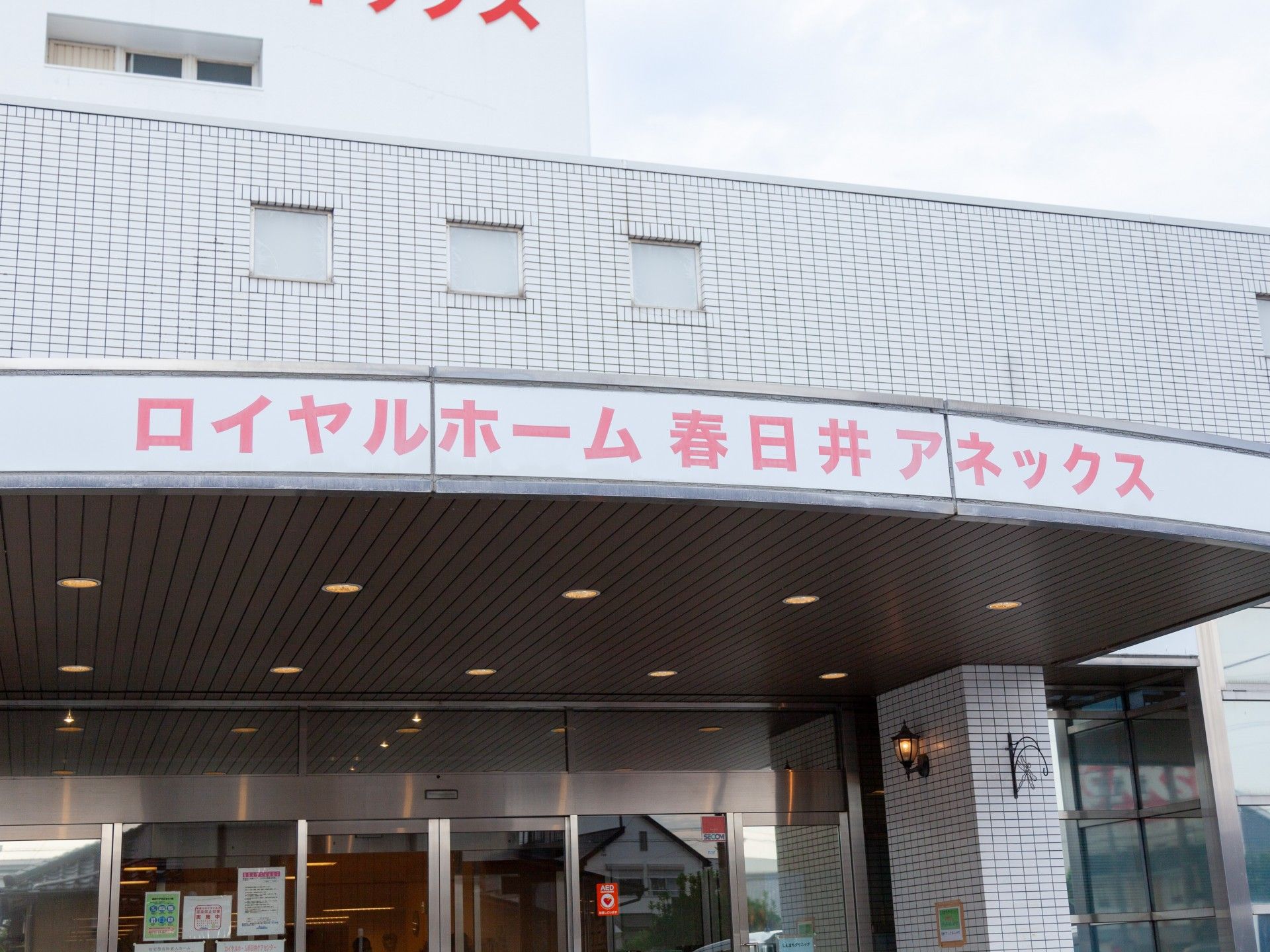 白を基調とした建物の玄関には屋根が付き、外壁には赤い文字でホーム名が書かれ、敷地内にある駐車場には車が止まっている。