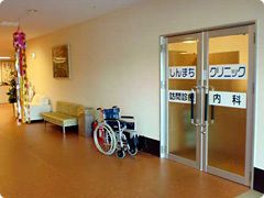 車いすが置かれた廊下にはガラス戸が設置されていて「しんまちクリニック」と書かれている。壁にはソファや消火栓もある。