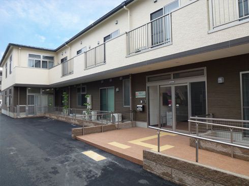 駐車場やベランダが付いた２階建ての建物。窓がたくさんつき、１階部分の出入り口には手すり付きのスロープが付いている。