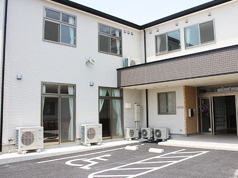 白を基調とした2階建ての建物。玄関先には屋根が付き、広い駐車場もあり、明るい雰囲気。