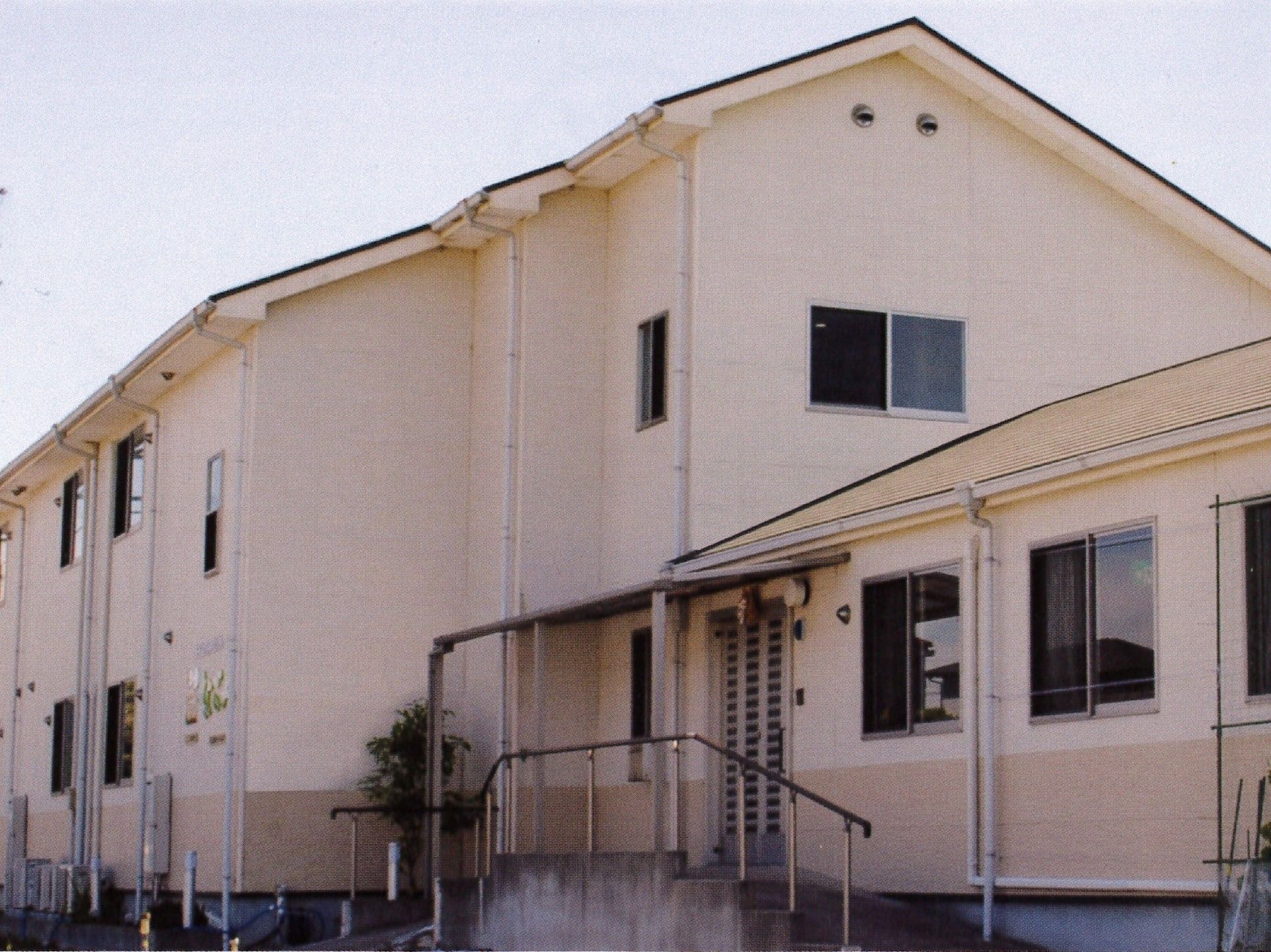 ２階建てのベージュの建物。三角屋根で、玄関には手すり付きの階段が付いている。