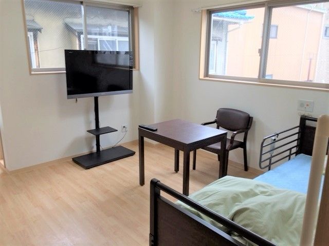 寝具付きのベッドが置かれたフローリングの部屋にはテレビやテーブルセットもあり、窓が2か所に付いている。