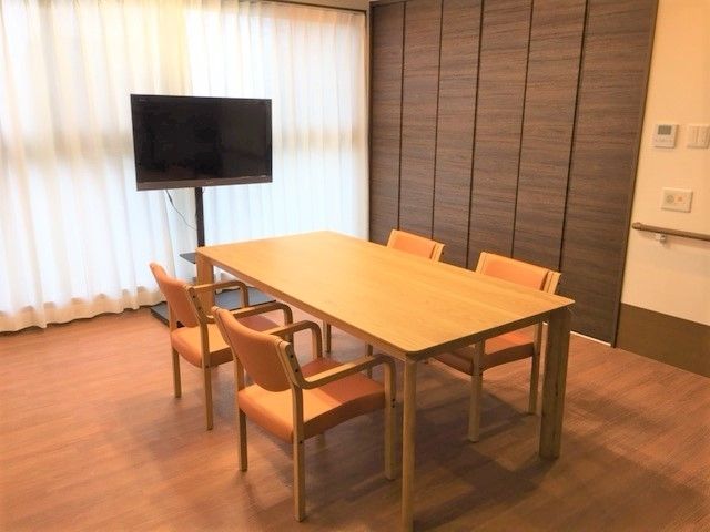 4人掛けのテーブルセットが中央に置かれた共有スペース。窓にはレースカーテンとカーテンが付き、テレビも置かれている。