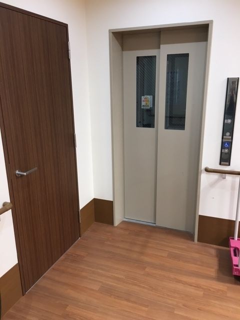 グレーの扉が設置されたエレベーター。周辺には何も置かれておらず、壁には手すりが設置され、操作ボタンが右側にある。