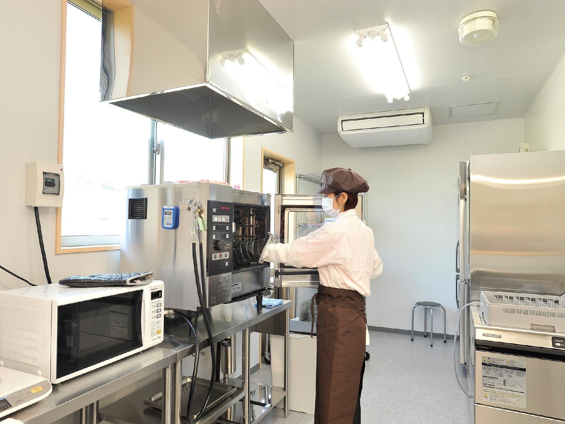 業務用のエアコンが設置された広い厨房には本格的な調理器具がそろっていて、白いシャツに茶色い帽子とエプロンを身につけたスタッフが作業をしている。