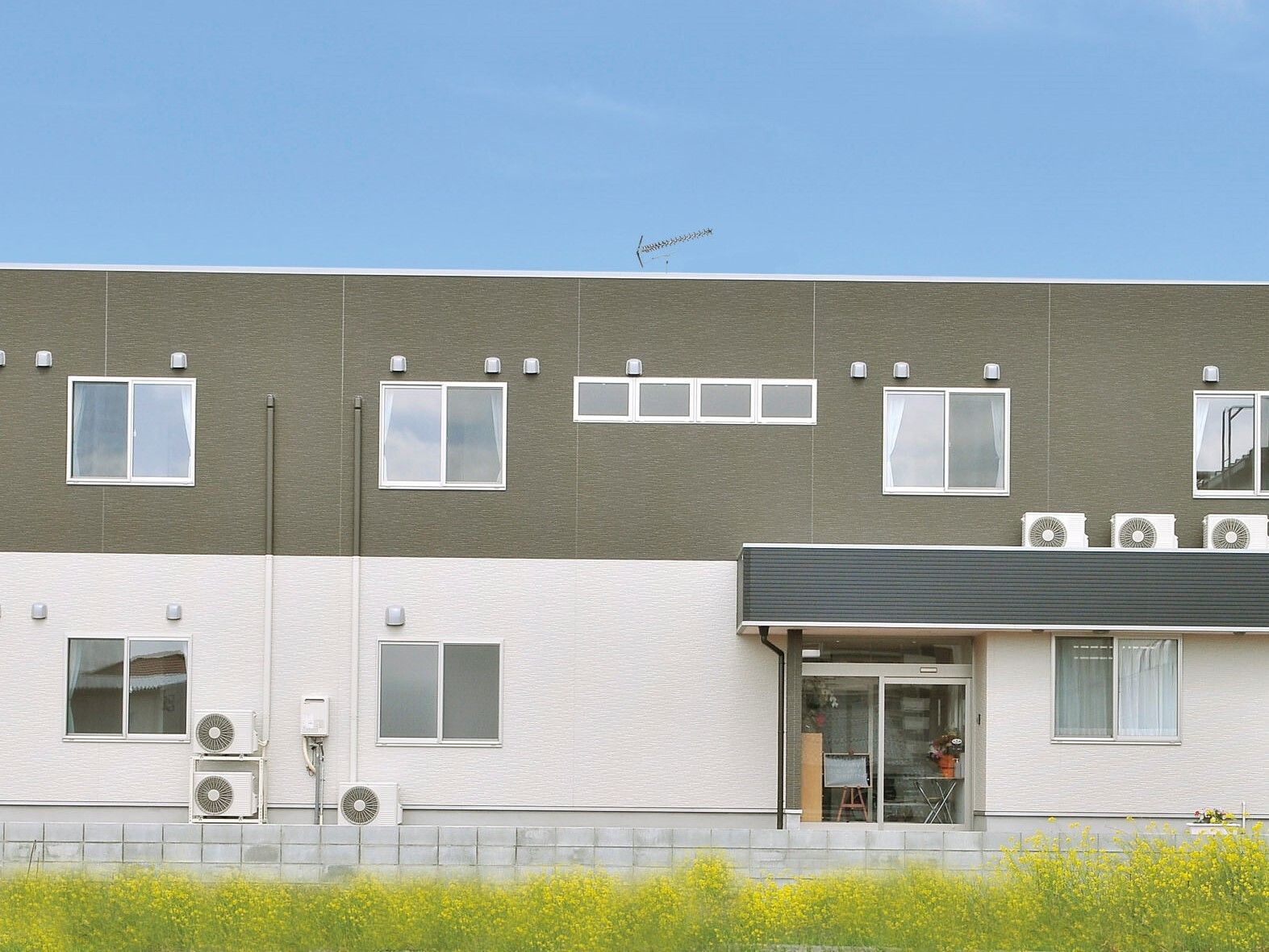 2階部分がグレーで、1階部分が白とモノトーンカラーでまとめられた2階建ての建物は横に長く、周りには緑が広がっている。