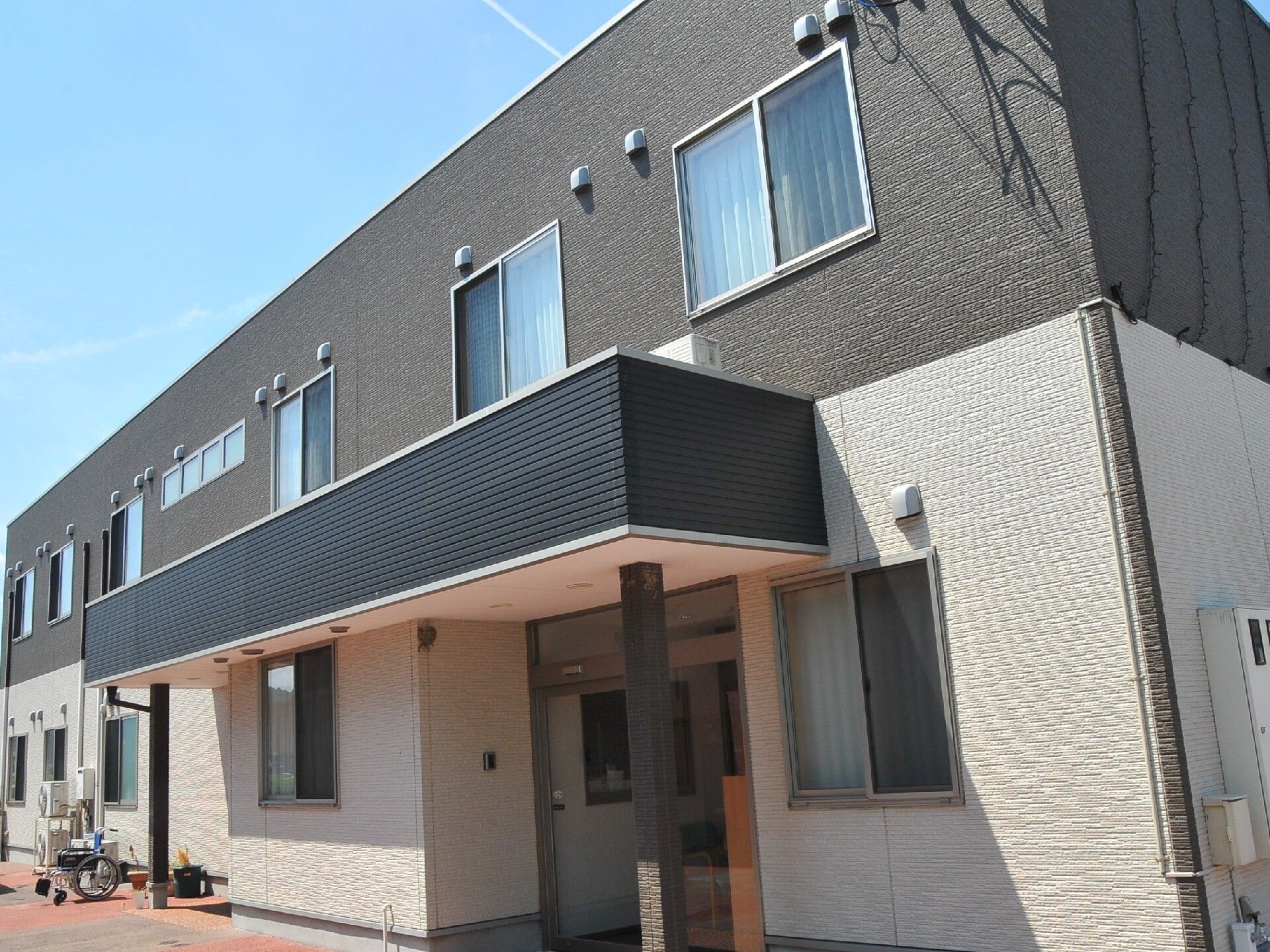 2階建ての建物は2階部分がグレーで1階部分が白。横に長く、玄関先は屋根が付いている。花が植えられたプランターも置かれている。