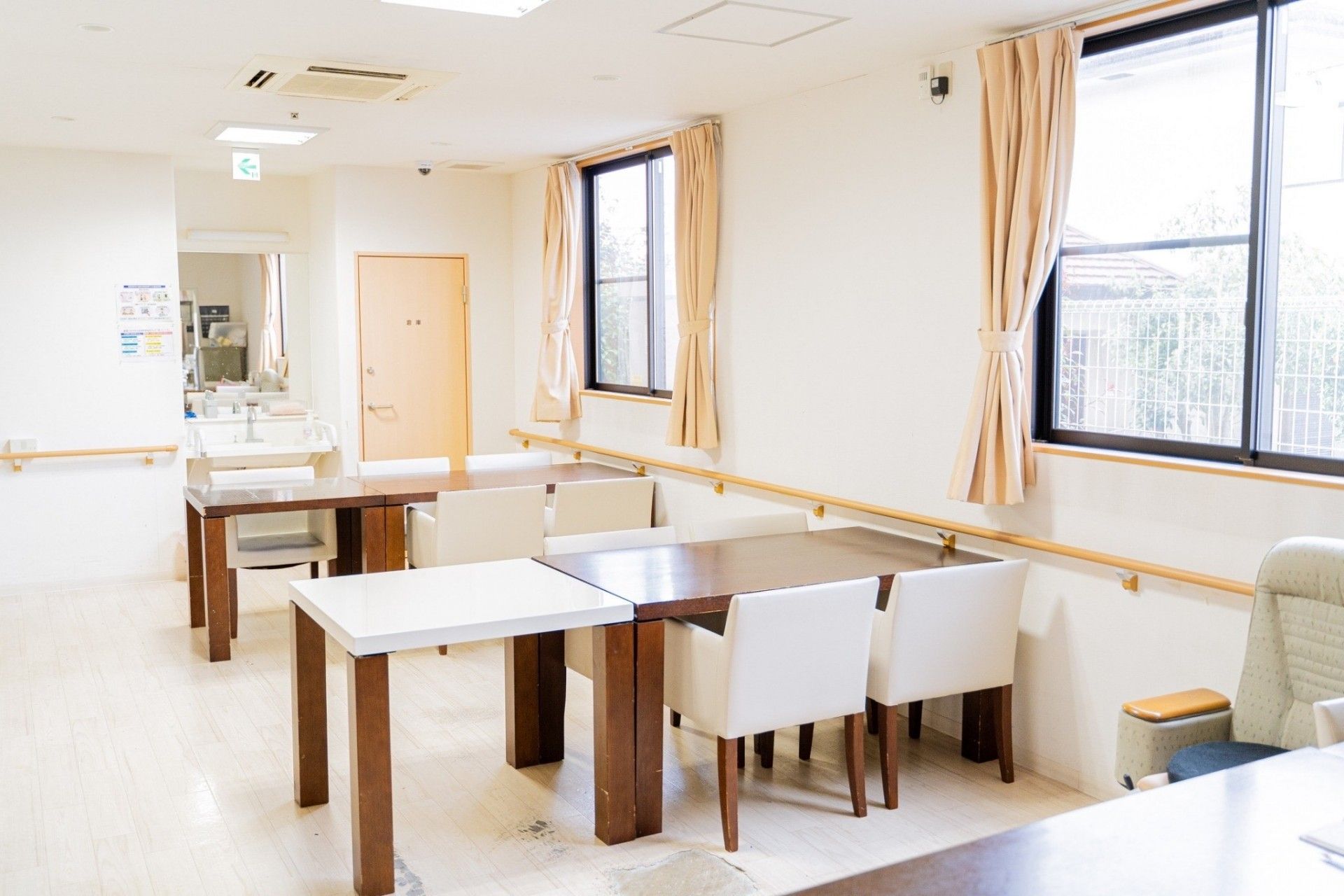 車いすの入居者も一緒に食卓を囲んでいる広々とした食堂。大きな窓がたくさんつき、明るい。窓のカーテンは1つを除き閉められている。