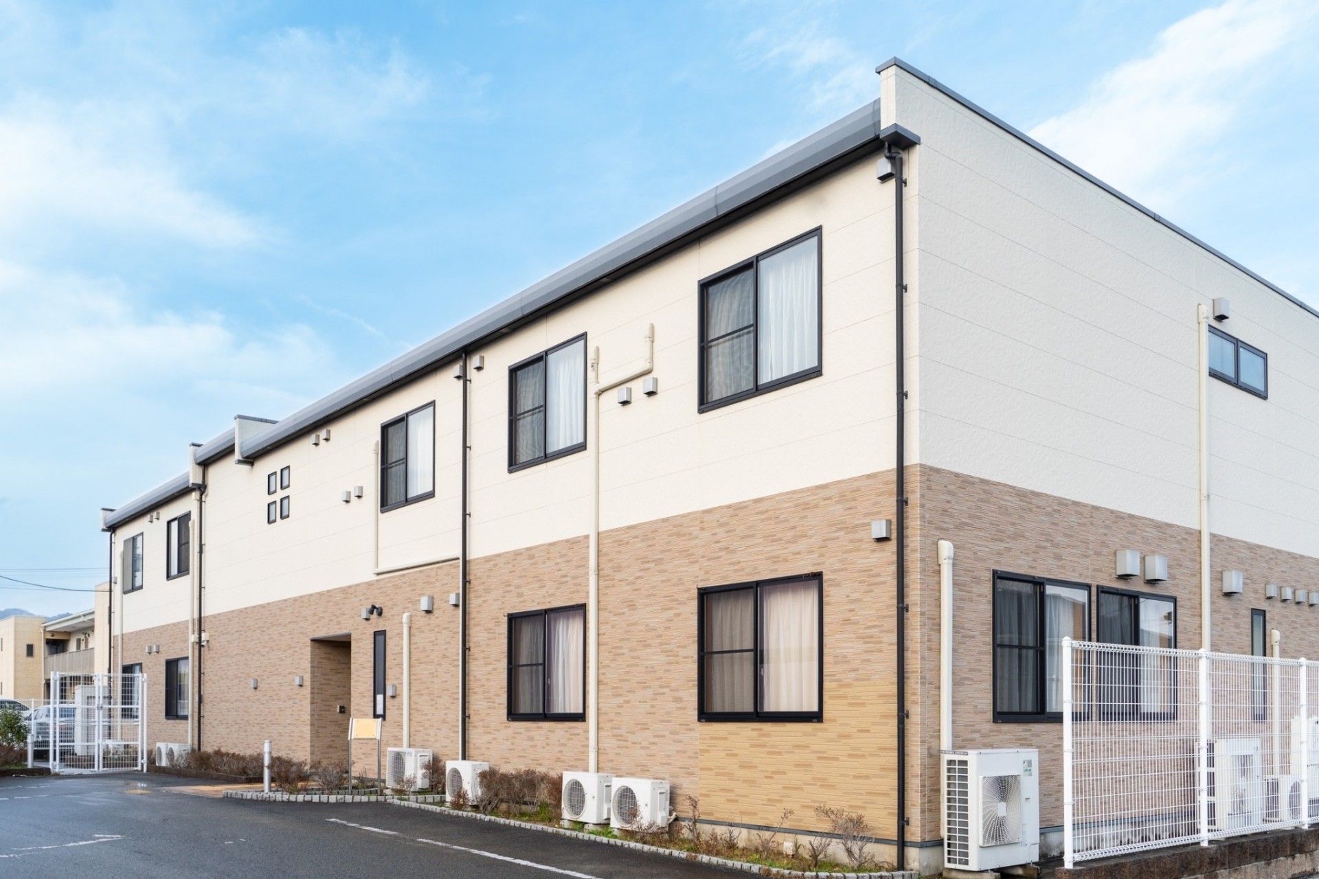 2階部分が白、1階部分がベージュの2階建ての建物。建物の周りには白いフェンスが建てられ、低木も植えられている。