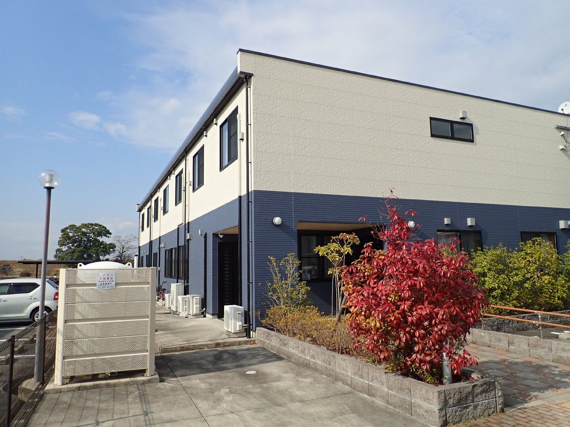 白とグレーの2階建ての建物の周りには緑が多く、広い駐車場もある。玄関には手すり付きのスロープがあり、男性が数名歩いている。