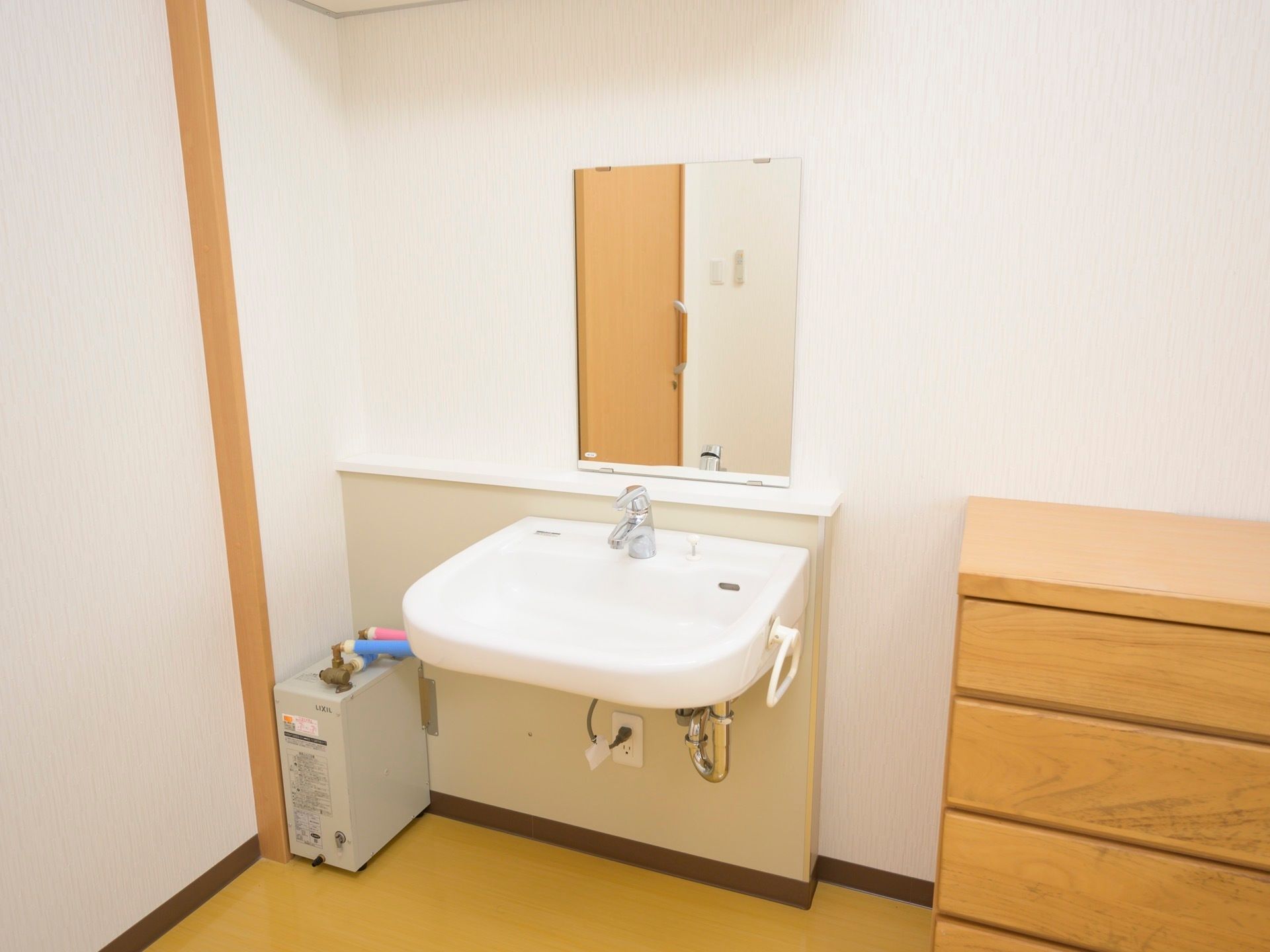 居室入口すぐの所に洗面台が設置されている。近くの床に置かれた灰色の器具に、水色とピンク色の配管がつながっている。