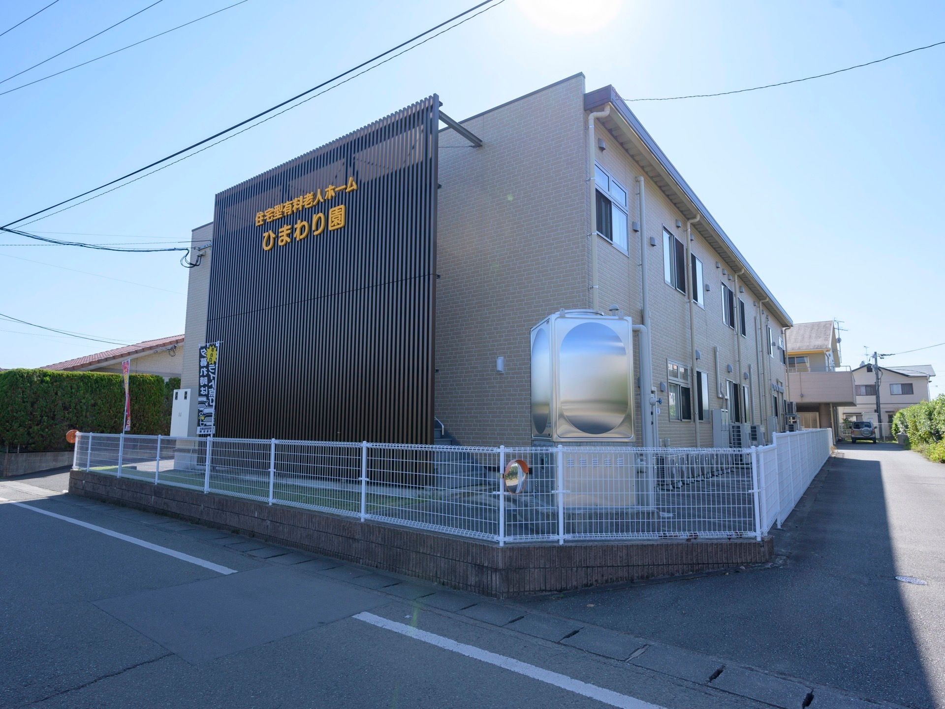 建物の周囲は白いフェンスで囲まれている。道路に面した格子状の外壁に大きな黄色の文字で施設名称が表示されている。