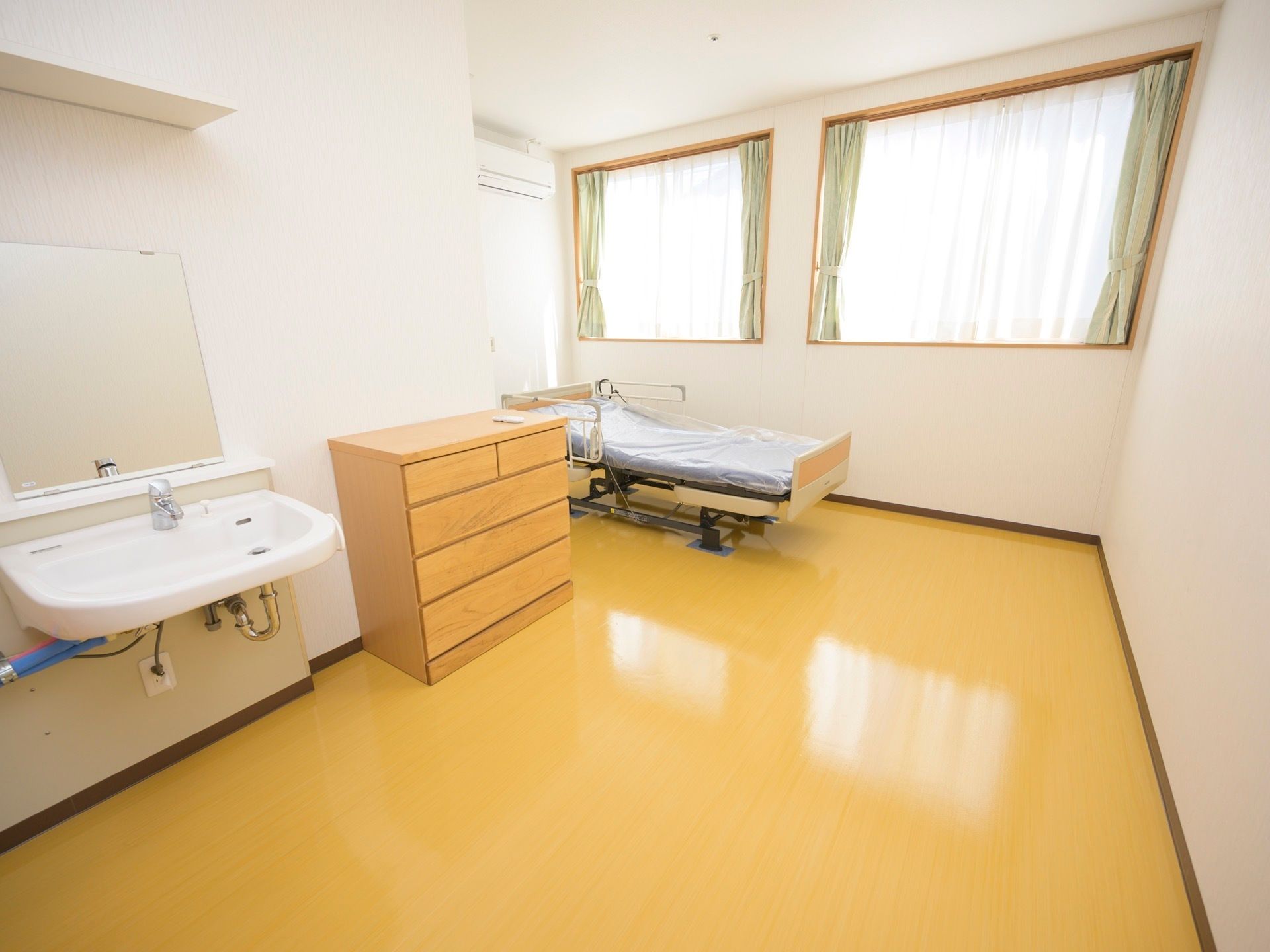 居室に腰高の窓が２つ並んで設置されている。介護ベッドの近くの壁の上部にエアコンが取り付けられている。