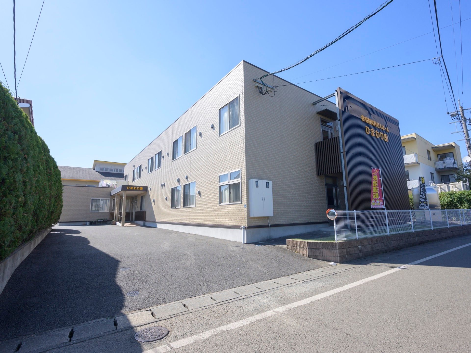 道路に面した敷地に建っている２階建ての施設である。敷地境界は背の高い木が並んで生垣のようになっている。