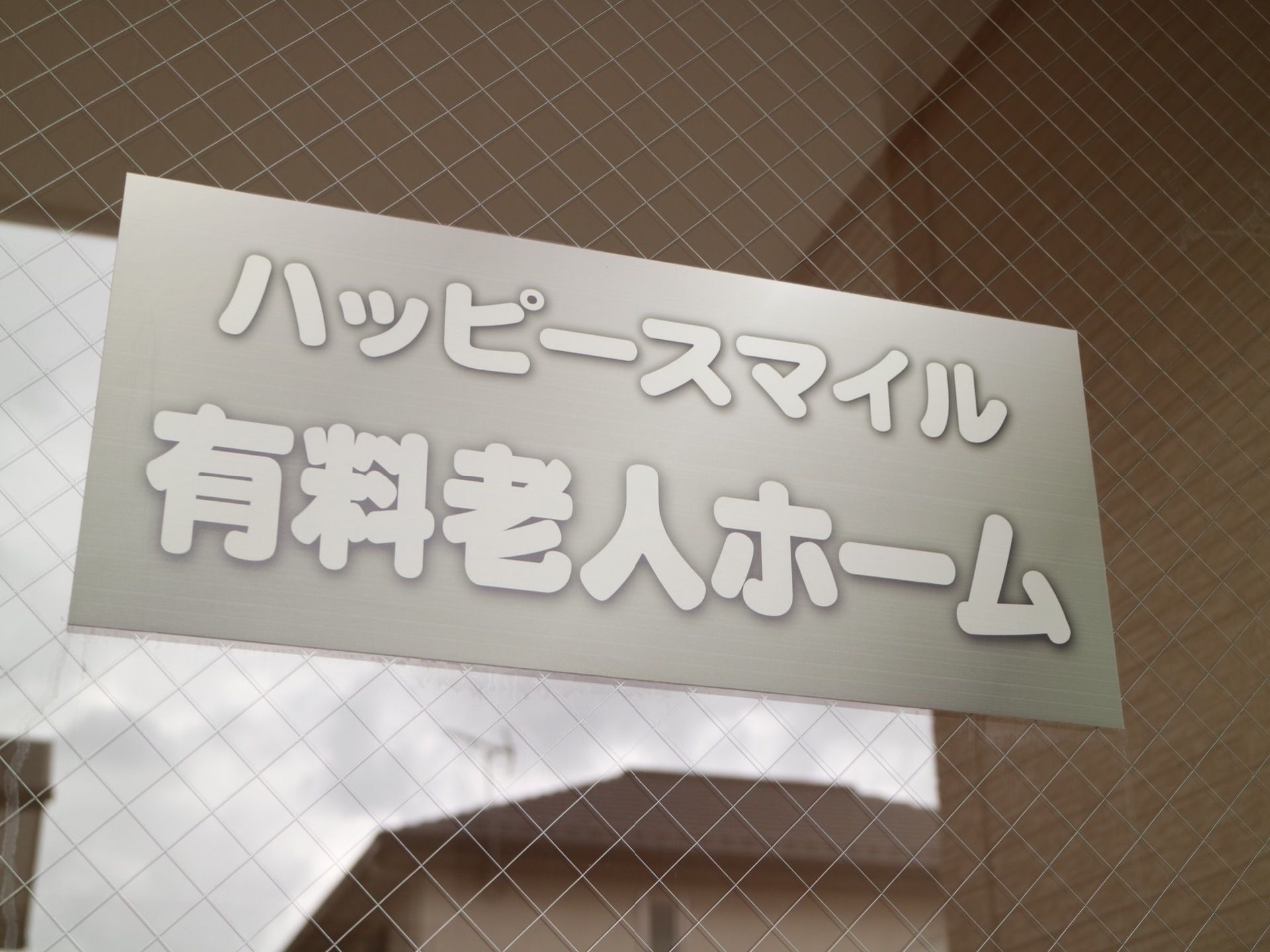 施設入口はガラス張りの自動ドア設置されており、施設名が書かれている。扉に反射して、向かい側に住宅がある。