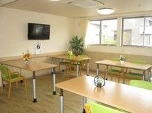 黄緑色のいすが添えられた木製のダイニングテーブルが複数並んだ食堂には、壁掛けテレビも設置されている。
