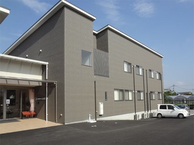 玄関周りには段差がなく、自動ドアで安全に出入りできる。建物はグレーの２階建てで、敷地内には広い駐車場も完備。
