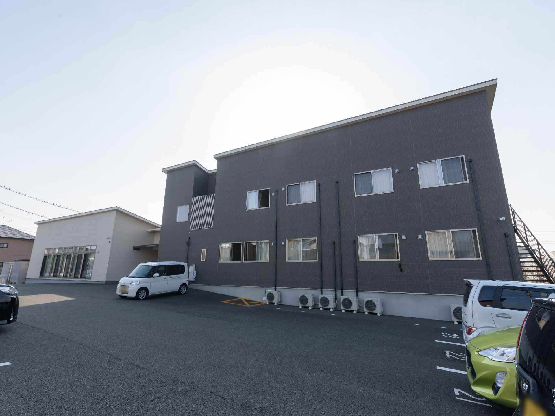 2階建てのグレーの外壁の建物で、外階段が設置されている。路面はアスファルトで舗装されており、車が数台停められている。