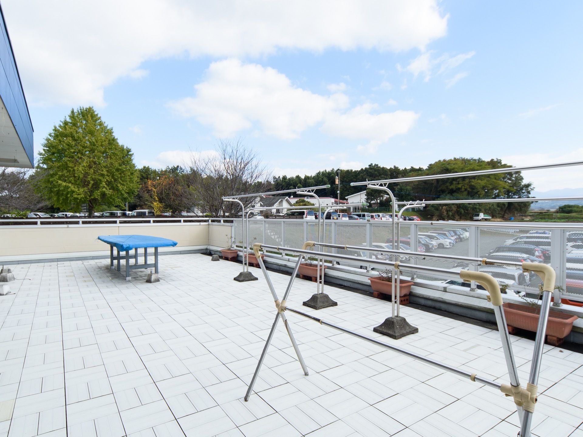 広々としたバルコニーに、大きい物干し台が２つと小さめの物干し台が１つ置かれている。バルコニーの周囲の柵の下には駐車場がある。