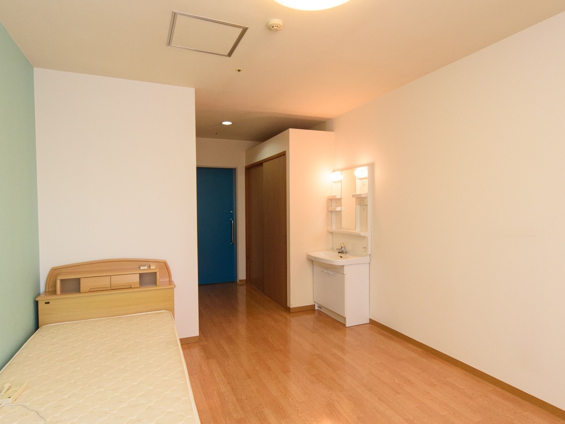 緑色の壁と白い壁に囲まれた洋風の居室。入口の扉は青色となっていて、白い洗面台と木目のベッドが置かれている。