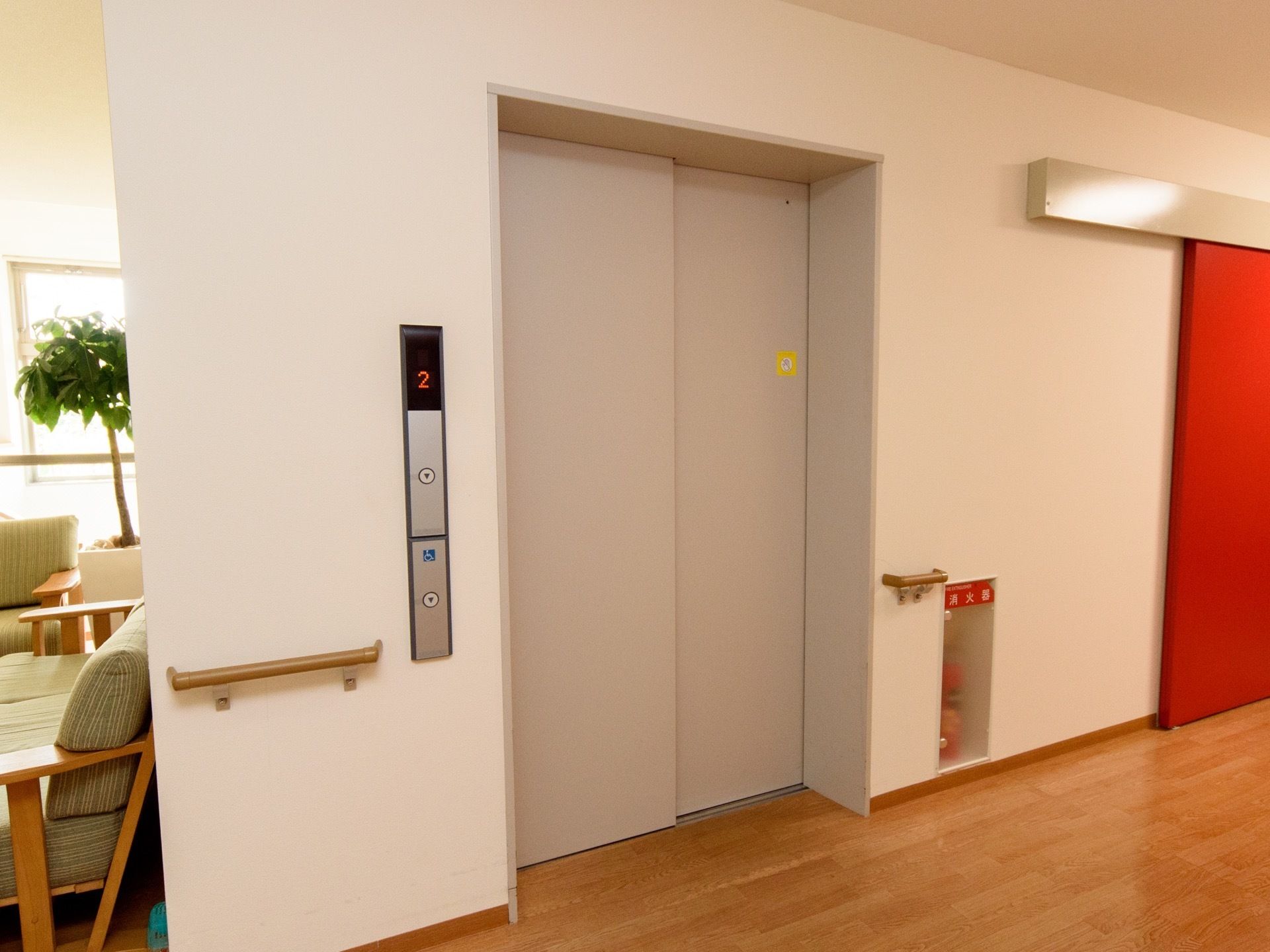 グレーのエレベーターが１基あり、その右には赤色の引き戸の扉がある。左側には緑色のソファ、観葉植物などが設置されている。