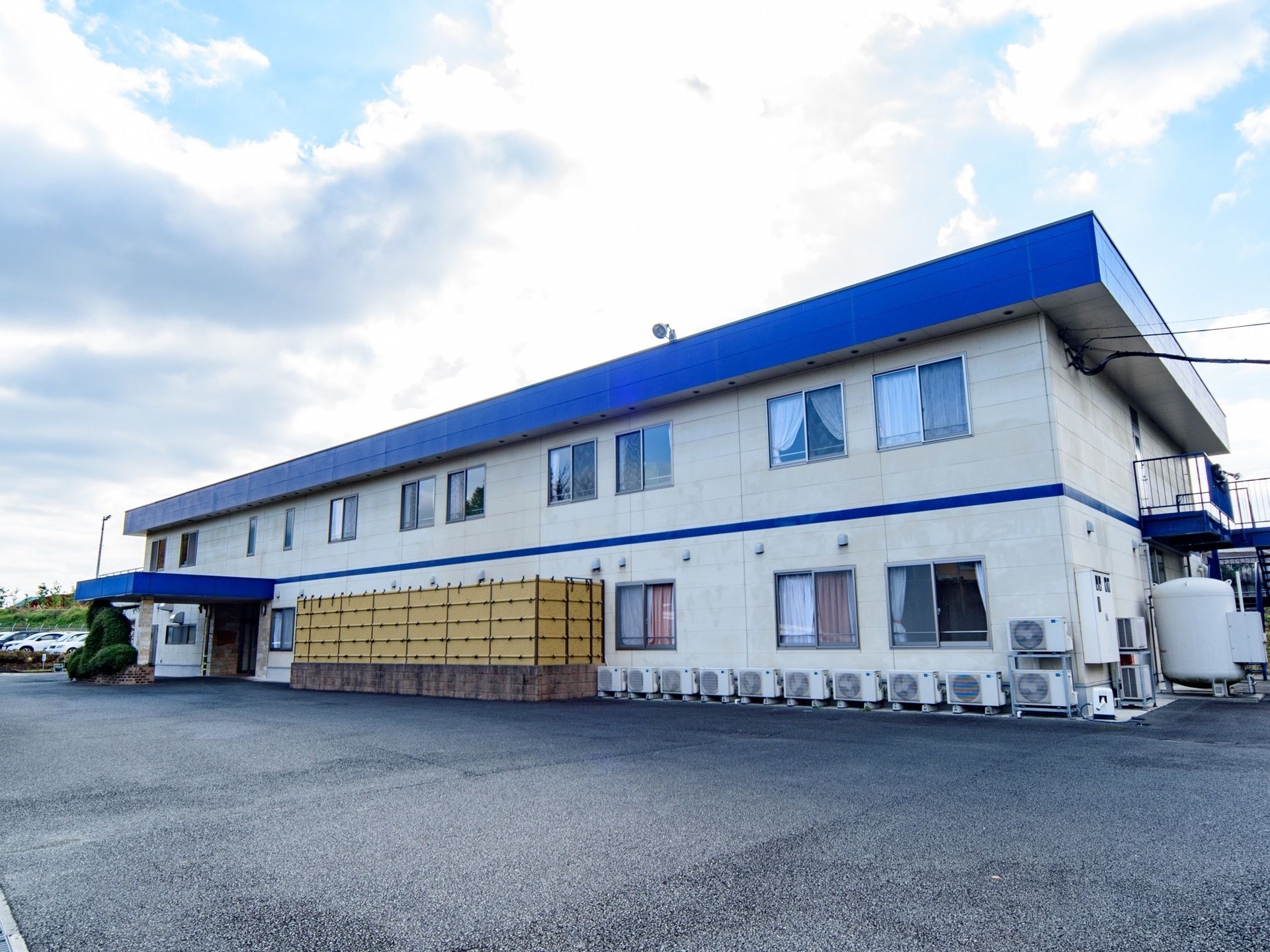 青色の屋根に白い壁をした２階建てのホーム。玄関の上には青色のひさしがあり、１階の一部の外壁は竹の垣根で囲まれている。
