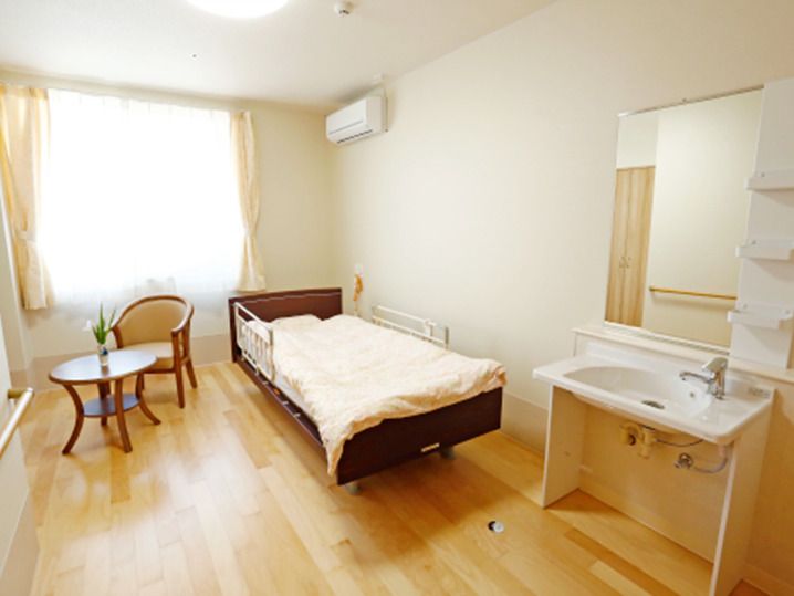 フローリングの部屋には寝具付きのベッドやテーブルセット、車いす対応の洗面所があり、大きな窓にはカーテンがある。