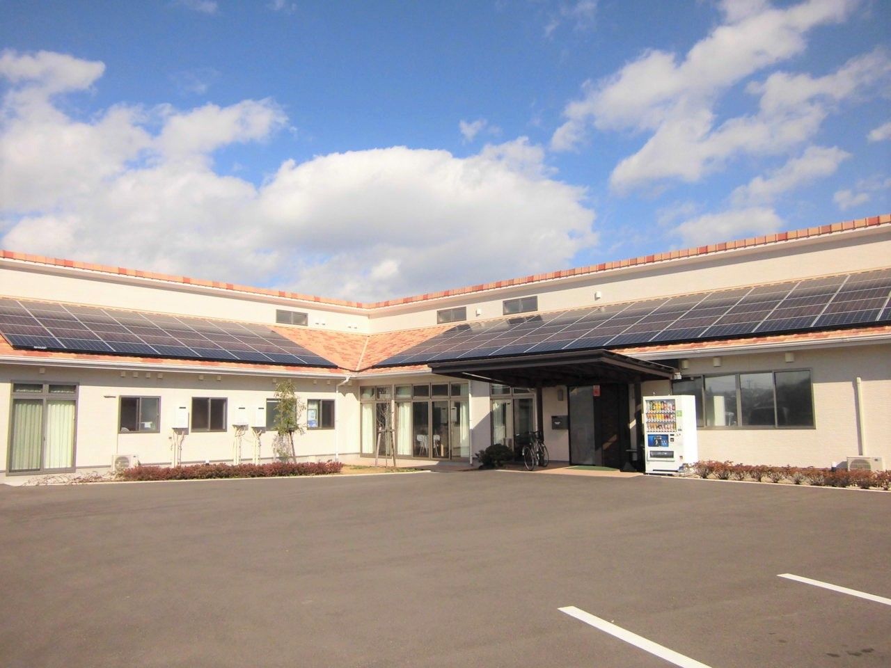 平屋建ての建物の瓦屋根にはソーラーパネルが設置され、広い駐車スペースもある。