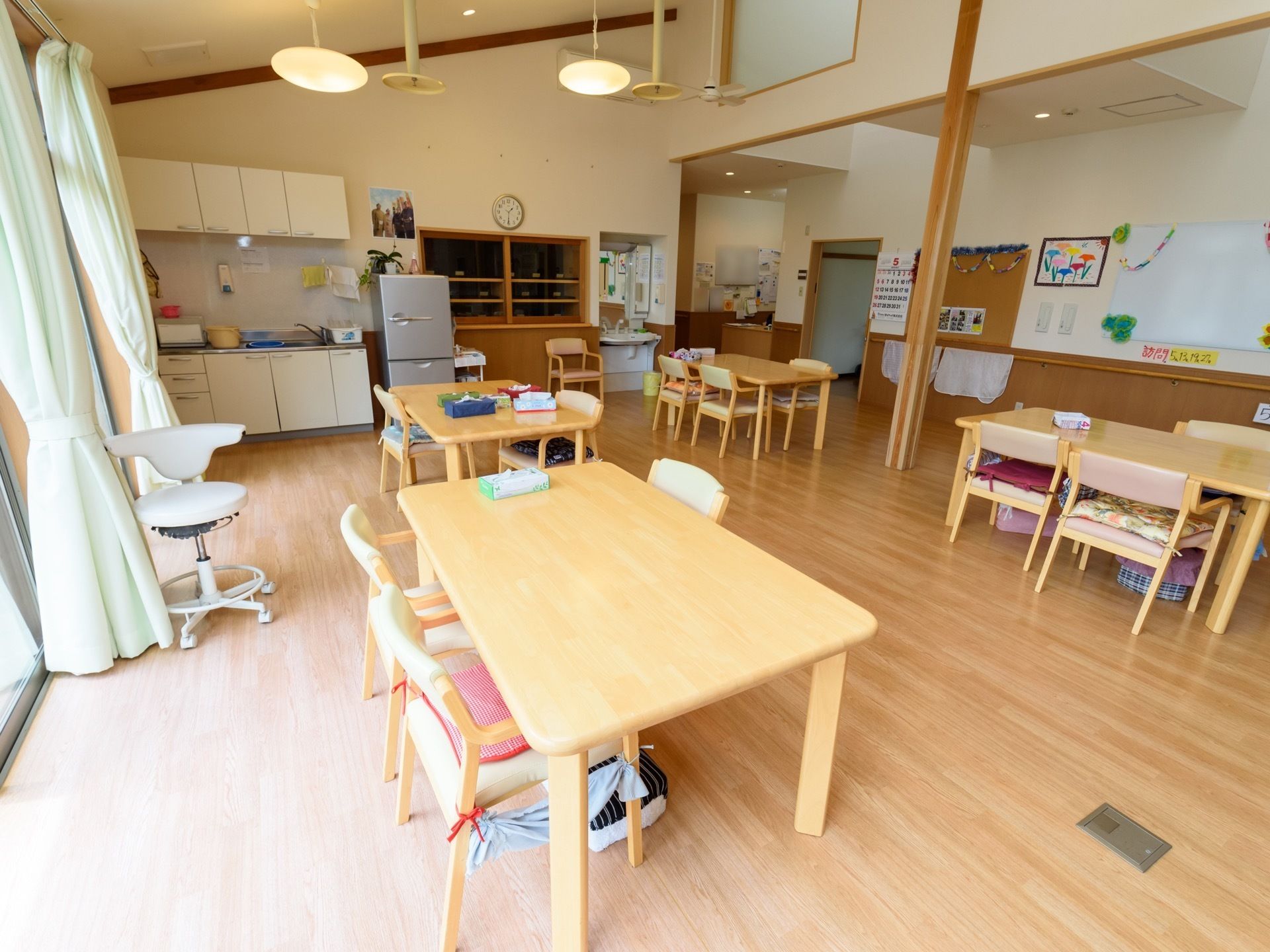 食堂に、テーブルが４台と多くの椅子が置かれている。キッチンの右横に冷蔵庫が設置されている。天井は斜めになっている。