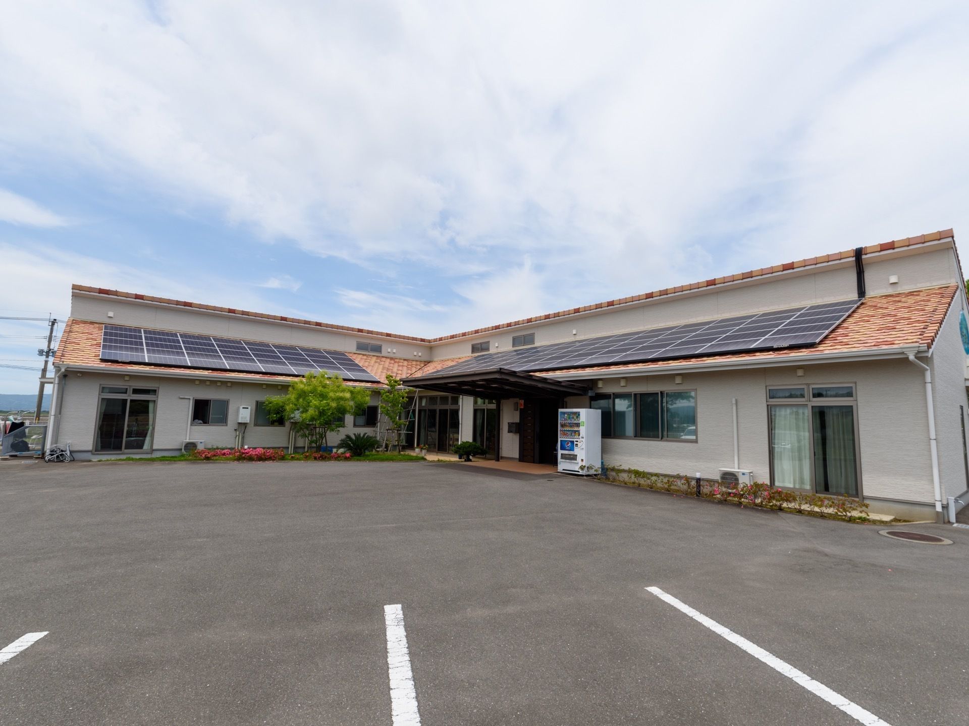 くの字型の構造をした平屋建ての施設である。屋根の上に大きな太陽光パネルが設置されている。駐車スペースの仕切りが描かれている。