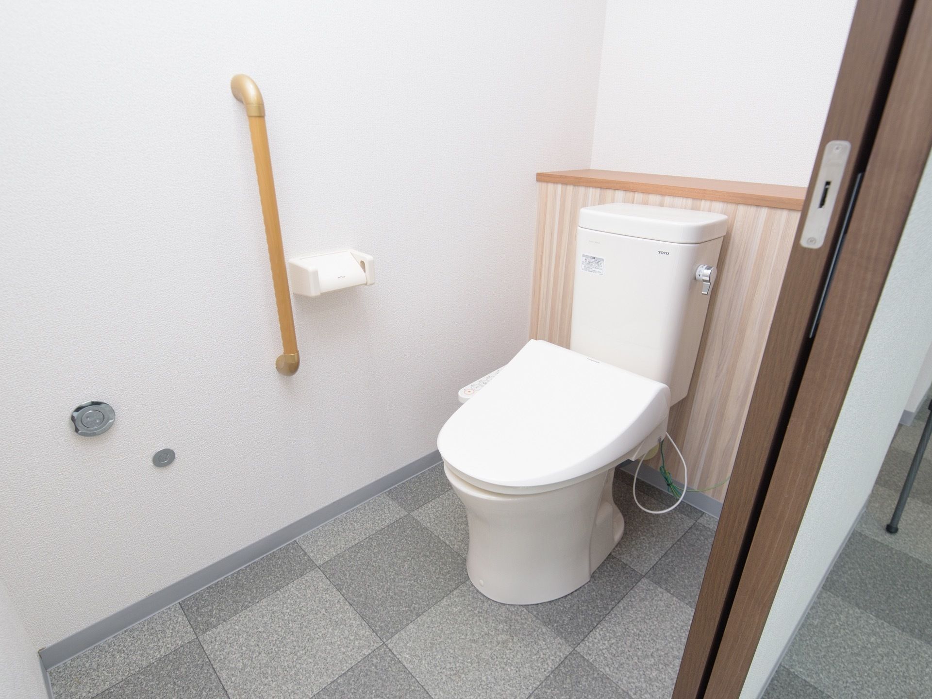 トイレは、洗浄機能付き便座を設置し、壁面にはペーパーホルダーの横に手すりを取り付けている。床はグレーの濃淡の格子柄である。