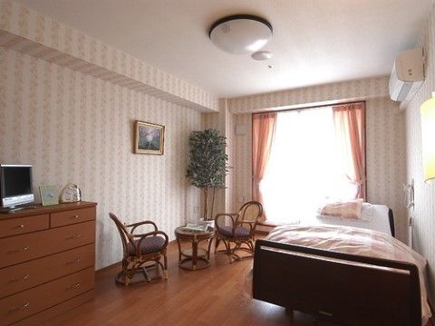 居室には大きなガラス戸があり、柄入りのクロスの雰囲気に合わせてレースとオレンジのカーテンを掛けている。天井にエアコンを完備している。