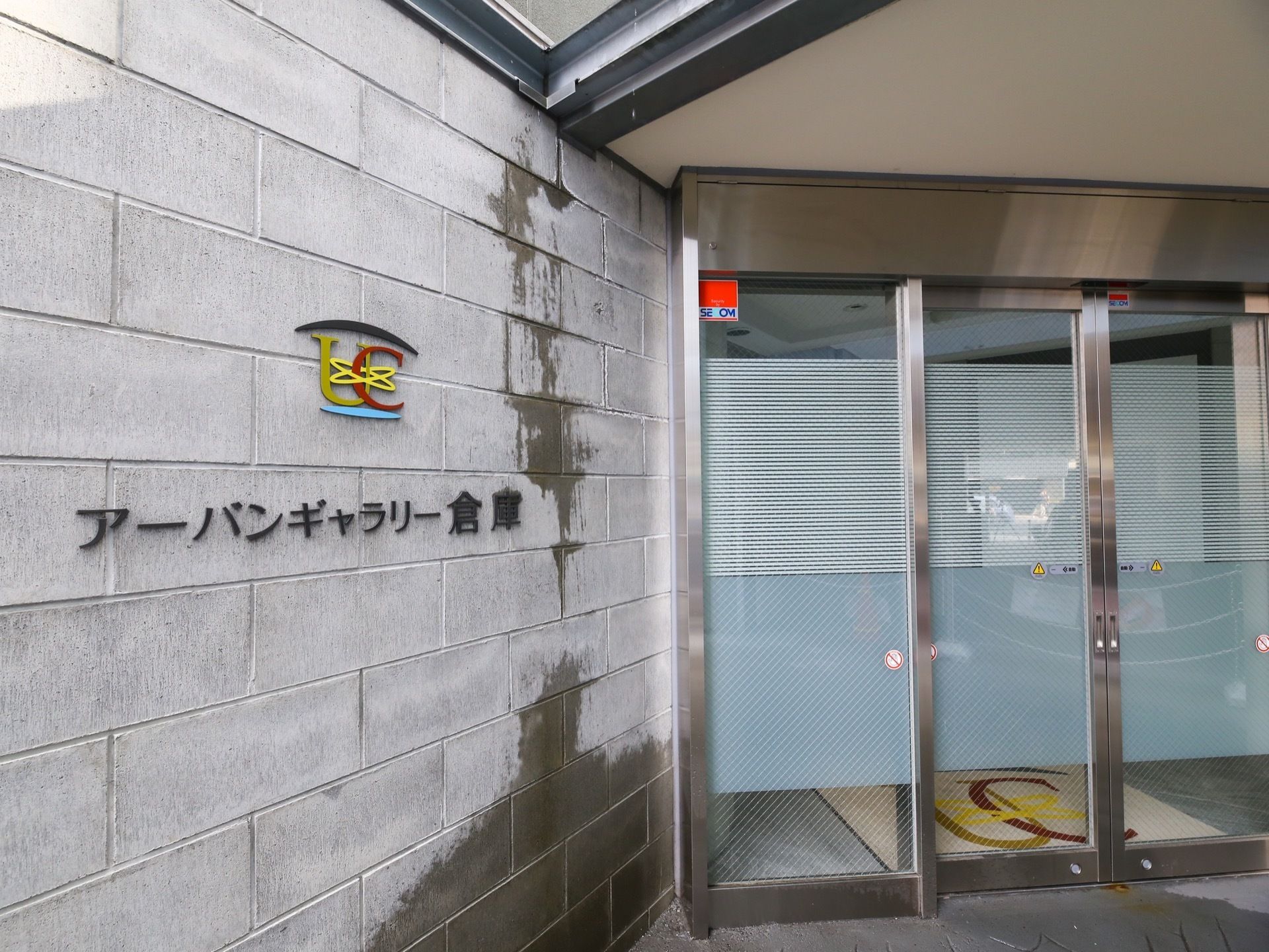 玄関は両開きの自動扉になっている。左上に警備会社のロゴマークのシールが貼られている。左側の壁に、施設のロゴと名称が表示されている。