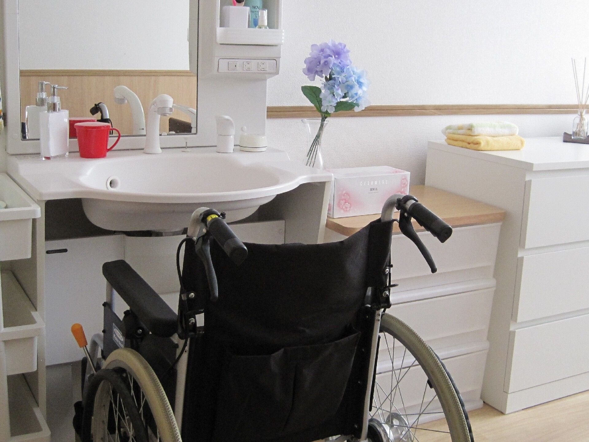 白いプラスチック製のチェストが複数置かれたフローリングの居室。車いすが洗面所にセットされ、洗面所回りにはソープやコップも置かれている。