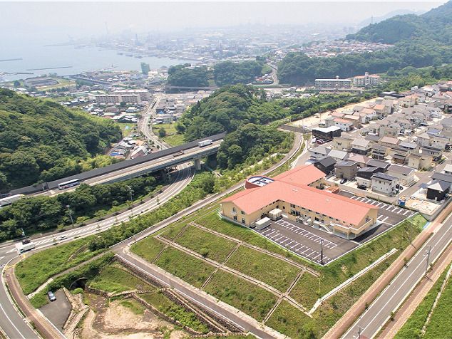 見晴らしのいい小高い丘にある赤い屋根の建物。敷地内には広い駐車場もあり、周りには住宅地があり、緑も多く、遠くには海も見える。