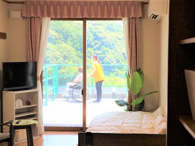 観葉植物やテレビ、棚、寝具付きのベッドなどが置かれた家庭的な雰囲気の部屋。窓の外に広がるウッドデッキには女性スタッフに付き添われた車いすの女性がいる。