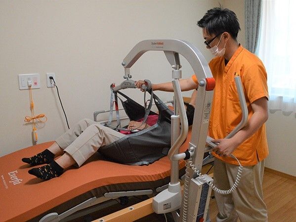 オレンジのユニフォームを着た眼鏡姿の男性スタッフがい畳用リフトを使用して介護ベッドに寝ている人を移動しようとしている様子。