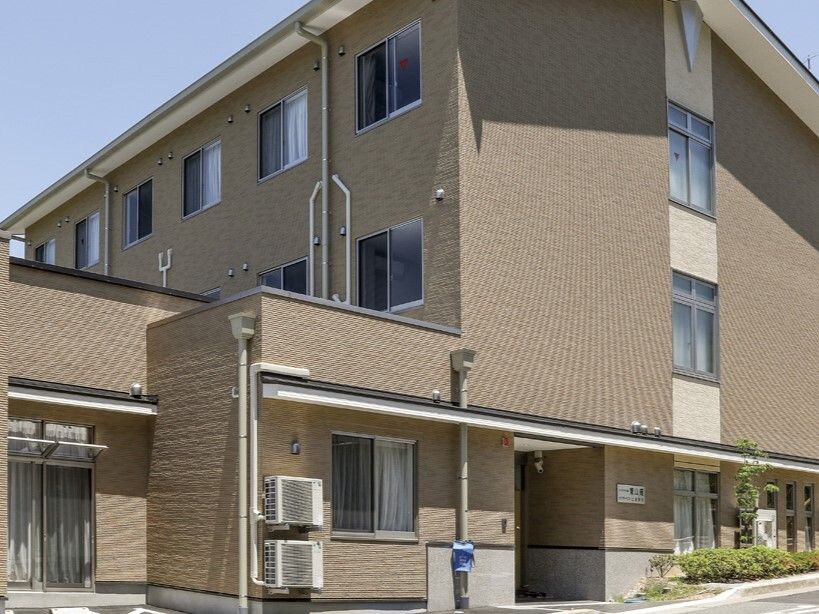 ブラウン系の建物は3階建てで、周囲には草木が植えられ、玄関先の壁には表札がある。